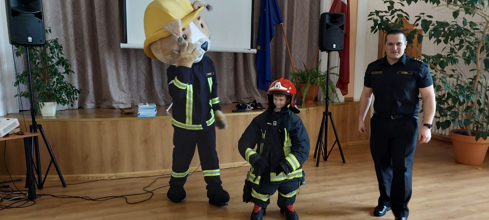 Zālē pie skatuves stāv bērns, kas ietērpts ugunsdzēsēja glābēja tērpā. Bērnam kreisajā malā stāv ugunsdzēsējs glābējs Guntiņš un skatās uz bērnu. Bērnam labajā pusē stāv VUGD nodarbinātais un skatās fotogrāfa virzienā