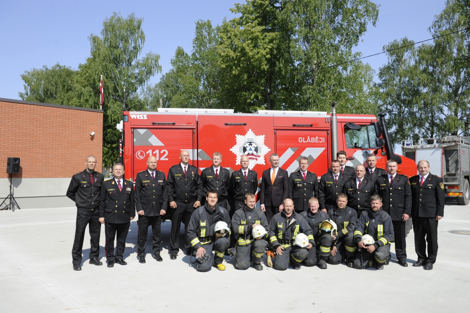 VUGD nodarbināto un iekšlietu ministrijas pārstāvja kopbilde. Priekšplānā pietupušies seši ugunsdzēsēji glābēji, kuri rokās tur baltās aizsargķiveres. Fonā citi VUGD nodarbinātie un iekšlietu ministrijas pārstāvis. Fonā sarkanā autocisterna