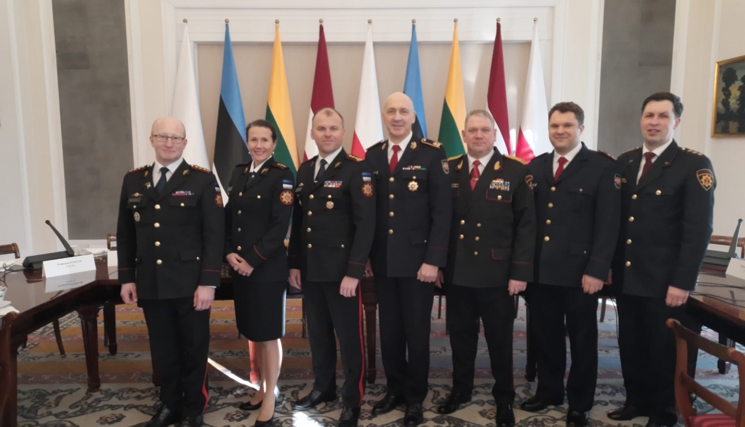 Septiņi konferences dalībnieki, tērpušies savās dienestu formās, stāv pie konferences dalībvalstu karogiem un skatās fotogrāfa virzienā