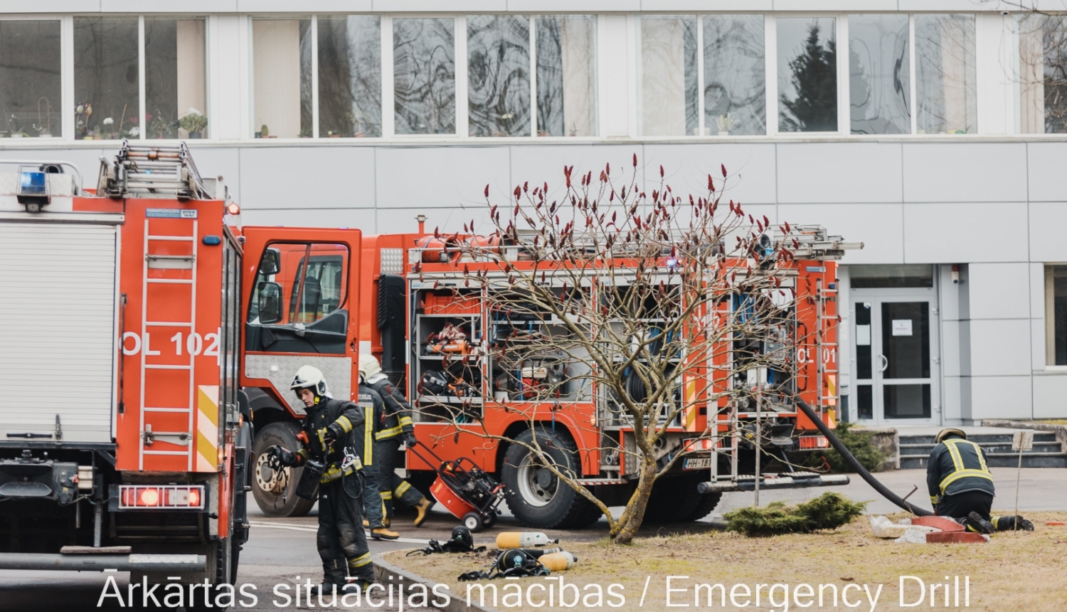 Divas Valsts ugunsdzēsības un glābšanas dienesta mašīnas stāv pie baltas ēkas. Ap mašīnām rosās ugunsdzēsēji glābēji.